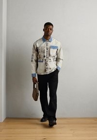 Leichte, gemusterte Bluse in Creme und Blau mit Kragen, Knopfverschluss und grafischen Designs. Kombiniert mit schwarzen Hosen und einer Tasche.