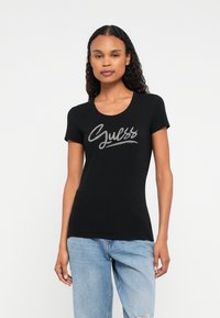 Kobieta z kręconymi włosami, ubrana w czarny dopasowany T-shirt z napisem "Guess" w srebrnym kolorze oraz jasnoniebieskie dżinsy z przetarciami, stojąca na jednolitym tle.