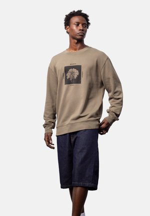 Uomo con felpa beige con stampa floreale e testo, abbinata a pantaloncini di jeans scuri fino al ginocchio, in piedi su uno sfondo semplice.