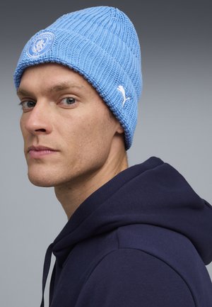 Light blue knitted beanie with a ribbed texture, featuring a circular Manchester City logo and a small white Puma emblem on the side.