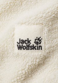 Krämfärgad fleeceväv med en strukturerad yta. Har en fyrkantig tygskylt med "Jack Wolfskin" och ett tassgrafik i svart.