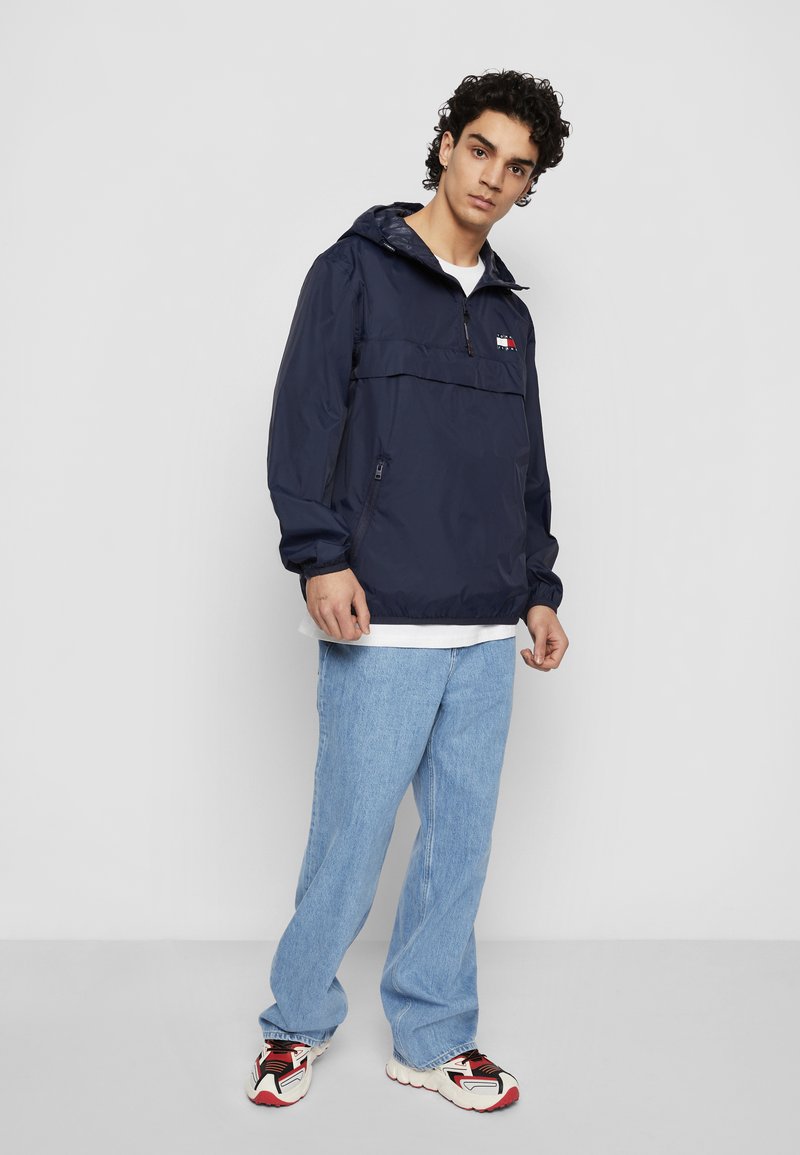 Navy windbreaker jacket with half-zip, elastic cuffs, and front pocket. Worn over a white shirt, paired with light blue jeans and patterned sneakers.