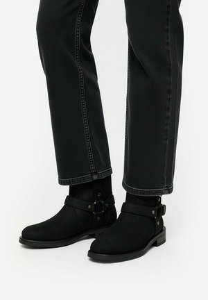 Stivaletti neri alla caviglia con cinturini e borchie metalliche indossati con jeans neri a gamba dritta su sfondo bianco.