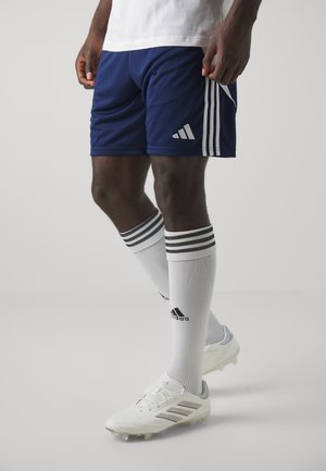 Man draagt marineblauwe Adidas-short, witte Adidas kniekousen met zwarte strepen en witte Adidas voetbalschoenen, staand op een vlakke vloer.