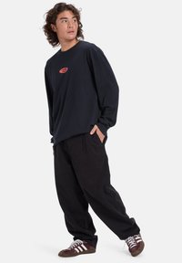 Schwarzes Langarmshirt mit rotem Logo, kombiniert mit lockeren schwarzen Hosen und braunen Sneakers mit weißen Streifen. Lässiger, entspannter Schnitt.