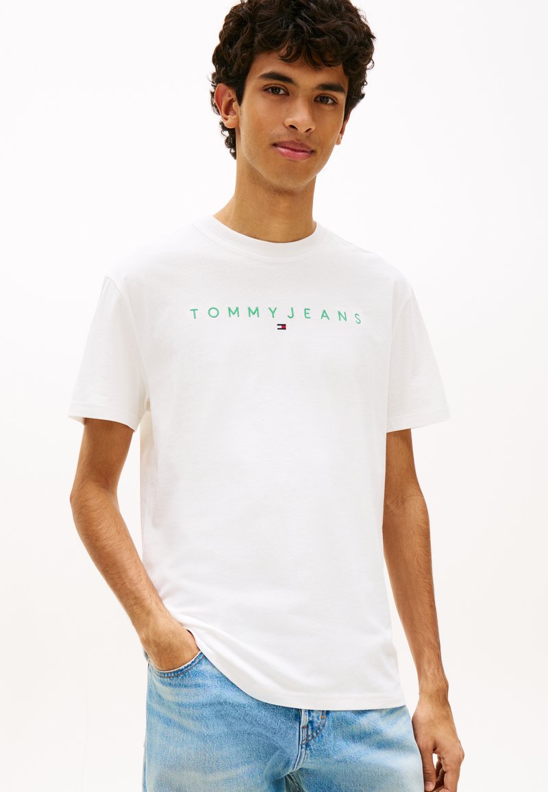 Νέος άνδρας που φοράει λευκό T-shirt Tommy Jeans και μπλε τζιν, με το ένα χέρι στην τσέπη, στέκεται μπροστά σε απλό φόντο.
