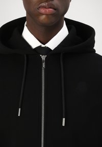 Sweatshirt preto com capuz, de textura suave, apresentando um fecho de correr frontal completo, acessórios prateados e um discreto detalhe de logótipo. Colarinho de camisa branca visível.