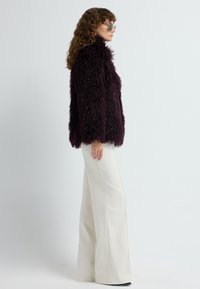 Kvinde med krøllet hår iført grønt farvede solbriller, en mørk lilla fuzzy jakke og hvide wide-leg bukser, som står i profil.