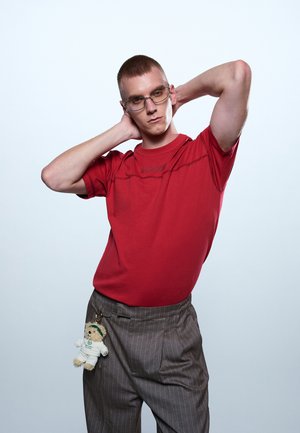 Νεαρός άνδρας με γυαλιά, κόκκινη μπλούζα και παντελόνι με λεπτές ρίγες, με ένα μικρό μπρελόκ αρκουδάκι, ποζάροντας με τα χέρια πίσω από το κεφάλι.