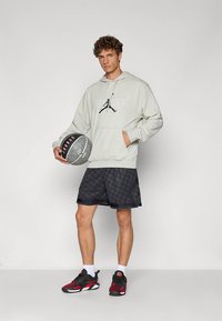 Camisola com capuz cinzenta clara com logótipo, calções pretos com padrão, sapatilhas desportivas pretas e vermelhas, e a segurar uma bola de basquetebol. Visual desportivo simples e casual.