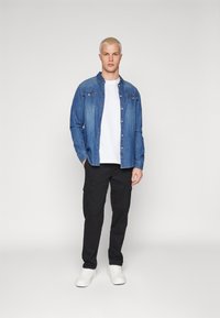 Cămașă din denim albastru peste un tricou alb, asociată cu pantaloni cargo negri și adidași albi. Prezintă detalii cu nasturi și buzunare.