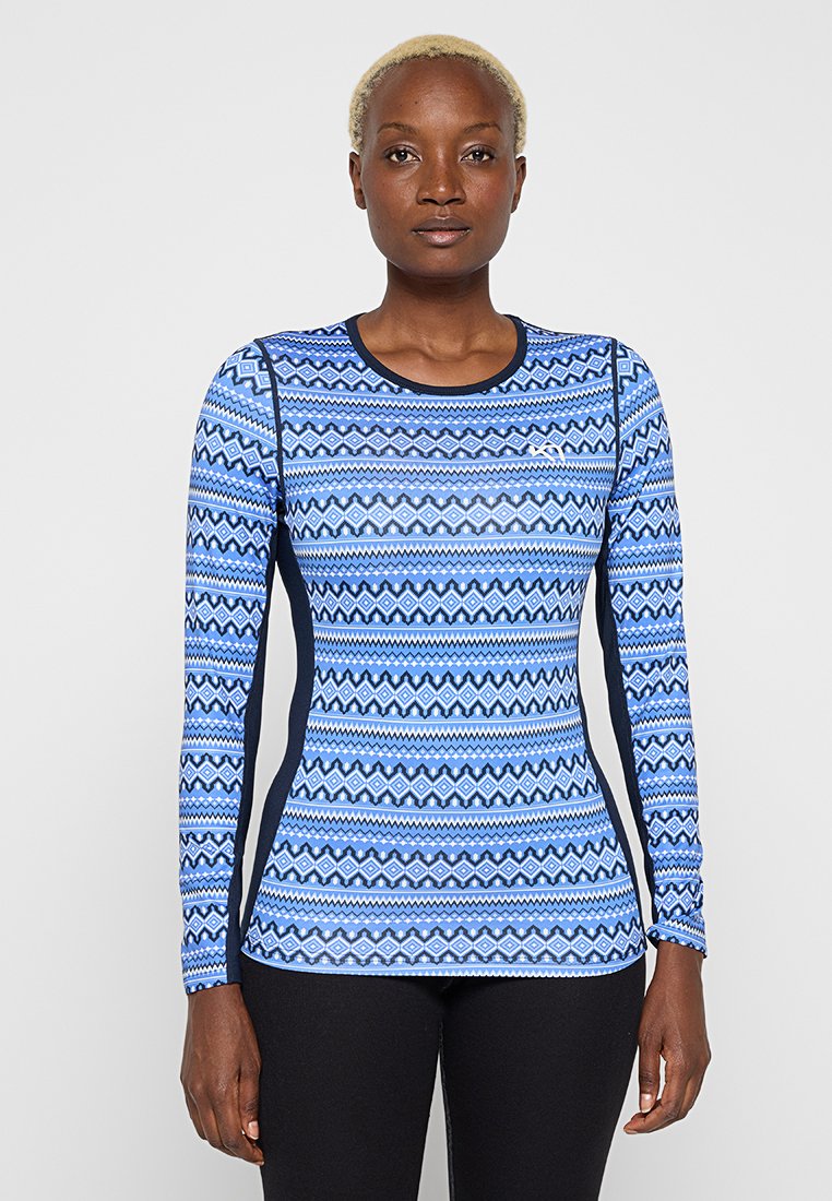 Femme portant un haut bleu ajusté à manches longues avec des motifs géométriques et des panneaux latéraux noirs, debout devant un fond uni.