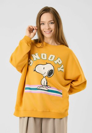 Junge Frau mit langen Haaren, die einen lockeren gelben Pullover mit Snoopy und bunten Streifen trägt, lächelt mit einer Hand nahe am Gesicht.