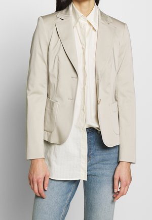 Woman wearing light beige tailored blazer over an off-white button-up shirt and light blue jeans, standing against a plain background.