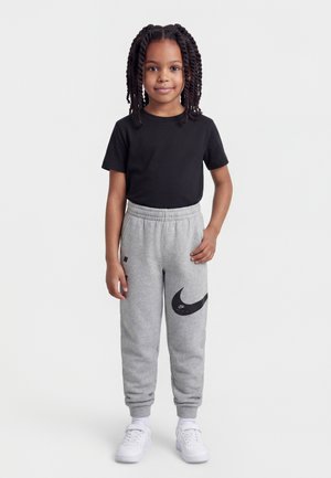 Fiatal lány áll, fekete pólót, szürke Nike melegítőnadrágot nagy Swoosh logóval, fehér tornacipőt és fehér zoknit visel, fehér háttér előtt.