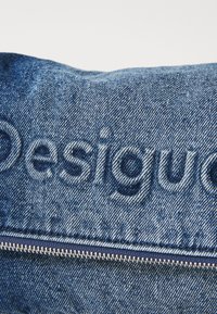 Denimová látka v modré barvě s vybledlým embosovaným logem "Desigual". Obsahuje stříbrný zip podél spodního okraje, který zdůrazňuje texturu a hloubku.