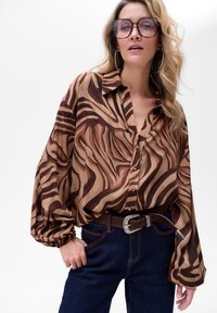 Bruine en crèmekleurige gestreepte blouse met losse mouwen, gecombineerd met donkere jeans en een bruine riem met een zilveren gesp. Duidelijke oversized brillen.