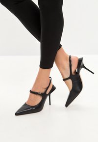 Tacchi slingback in pelle nera con punta affusolata e sottile tacco a stiletto. Presentano una cinghia con fibbia e lati tagliati per un ulteriore dettaglio di design.