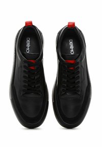 Baskets en cuir noir présentant une texture lisse, un bout rond, des lacets noirs et un détail de tirage rouge au niveau du talon. Notez la semelle en caoutchouc.