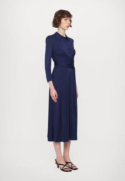 Haljina na preklop tamnoplave boje s ovratnikom, dugim rukavima, sprijeda je kopčana na gumbe i ima detalj s remenom u struku. Nosi se s crnim sandalama na petu s trakicama.