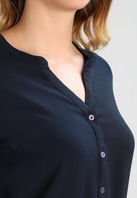 Marineblaue Bluse mit glatter Textur, ausgestattet mit einem dezenten V-Ausschnitt und kleinen lila Knöpfen entlang der Vorderblende.