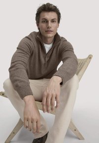 Brauner Zip-Up-Pullover mit strukturiertem Strick. Das Model trägt ein weißes Hemd darunter und helle beigen Hosen und sitzt auf einem Holzstuhl.