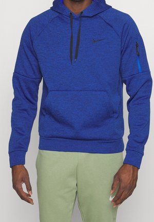 Mężczyzna ubrany w niebieską bluzę Nike z czarnymi ściągaczami i kieszenią na zamek na rękawie, w połączeniu z jasnooliwkowymi spodniami dresowymi Nike.
