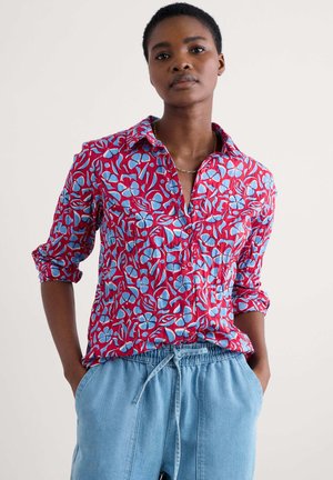 Red and blue patterned shirt with a button-down collar, rolled sleeves, and a relaxed fit. Worn with light blue drawstring pants.