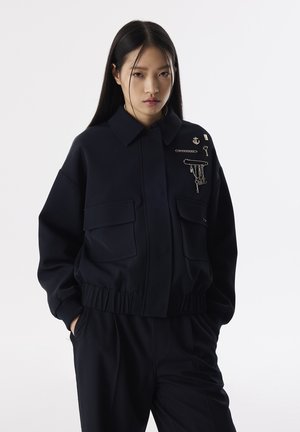 Navyblauwe cropped jas met een kraag, voorzien van twee grote zakken en decoratieve metalen pinnen, waaronder een anker en kettingen.