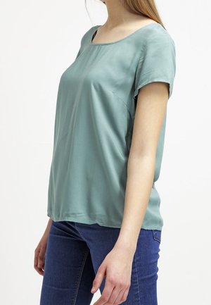 Femme portant un chemisier à manches courtes couleur teal clair et un jean bleu foncé, debout devant un fond blanc uni.