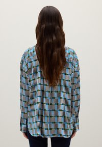 Person mit langen braunen Haaren, die ein gestreiftes und kariertes Langarmhemd in Blau, Grün, Schwarz und Weiß trägt, steht mit dem Rücken zu einem schlichten Hintergrund.