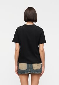 Femme aux cheveux courts et foncés portant un t-shirt noir uni et une jupe en jean courte avec de grandes poches arrière beige, debout devant un fond blanc.