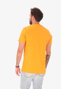 Man met tatoeages die een oranje T-shirt en lichtblauwe jeans draagt, staand met zijn rug naar de camera tegen een effen witte achtergrond.