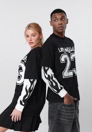 Doi tineri stau spate în spate purtând tricouri negre suprapuse cu mâneci albe cu flăcări și numărul 23 imprimat alb.