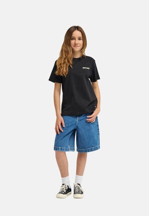 Jeune femme debout avec de longs cheveux, portant un t-shirt noir "element", un short en jean bleu, des chaussettes blanches et des baskets noires.