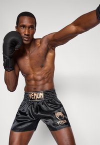Juodi satino bokso šortai su tekstūruota juosmens juosta, kurioje yra auksinis „Venum“ logotipas. Modelis dėvi juodas bokso pirštines ir pozuoja atletine poza.