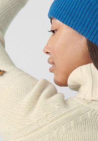 Maglione di colore crema a maglia con colletto alto e texture a coste, abbinato a un berretto a coste di un brillante blu. Cuciture dettagliate visibili.
