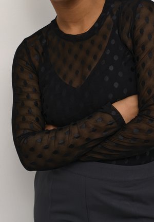 Personne portant un haut noir transparent à pois à manches longues avec un caraco noir en dessous, bras croisés et pantalon gris foncé.