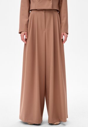 Wijde, hoog geplaatste bruine broek gecombineerd met een bijpassend cropped jasje, getoond van taille tot vloer met de handen van het model ontspannen langs het lichaam.