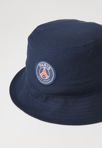 Tumes sinine päike müts, mis on valmistatud kergest kangast, esitleb ümmargust logot, millel on tekst "Paris Saint-Germain" ja dekoratiivne embleem.