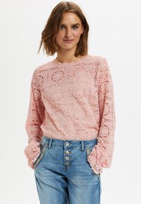 Top in pizzo a maniche lunghe di un delicato rosa, con intricate decorazioni floreali e un collo a scallop, abbinato a jeans in denim blu chiaro.