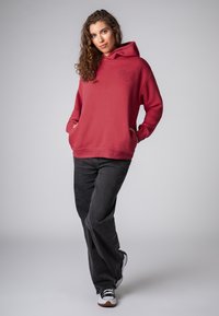 Rode hoodie met een voorzak en een hartjespatroon op de linkerborst, gecombineerd met zwarte wijde broekspijpen en zwart-witte sneakers.