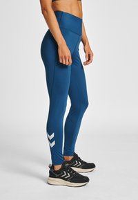 Kvinna st år i blå högmidjade leggins med vita chevronränder nära anklarna, bär svarta och vita sportskor.