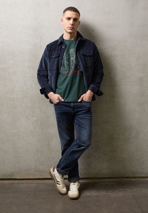 Giovane uomo con giacca blu scuro, t-shirt verde con grafica, jeans blu e sneaker bianche in piedi contro un muro di cemento grigio.