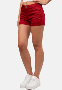 Shorts di denim rossi con una vestibilità slim, dotati di chiusura con bottone frontale e tasche anteriori. Indossati con sneakers bianche su uno sfondo neutro.