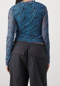 Långärmad topp i blå och svart zebramönster, gjord av genomskinligt material. Matchas med svarta cargobyxor med bakfickor.