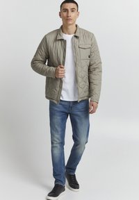 Quilted beige jakke med krage og glidelås, frontlomme, båret over en hvit skjorte, kombinert med blå jeans og svarte joggesko.