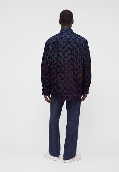 Homme debout, de dos, portant une chemise à manches longues sombre à motifs, un pantalon bleu et des baskets blanches sur un fond uni.