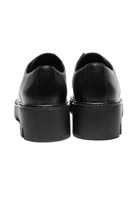 Musta nahkakengät pyöreällä muotoilulla, jossa on paksu, teksturoitu pohja ja minimaalinen ommel yläosassa. Takana näkyy sileä pinta.