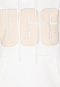 Biela mikina s kapucňou, ktorá má veľké, béžové logo UGG v mäkkej, textúrovanej výšivke. Obsahuje klokanie vrecko a nastaviteľnú šnúrku.